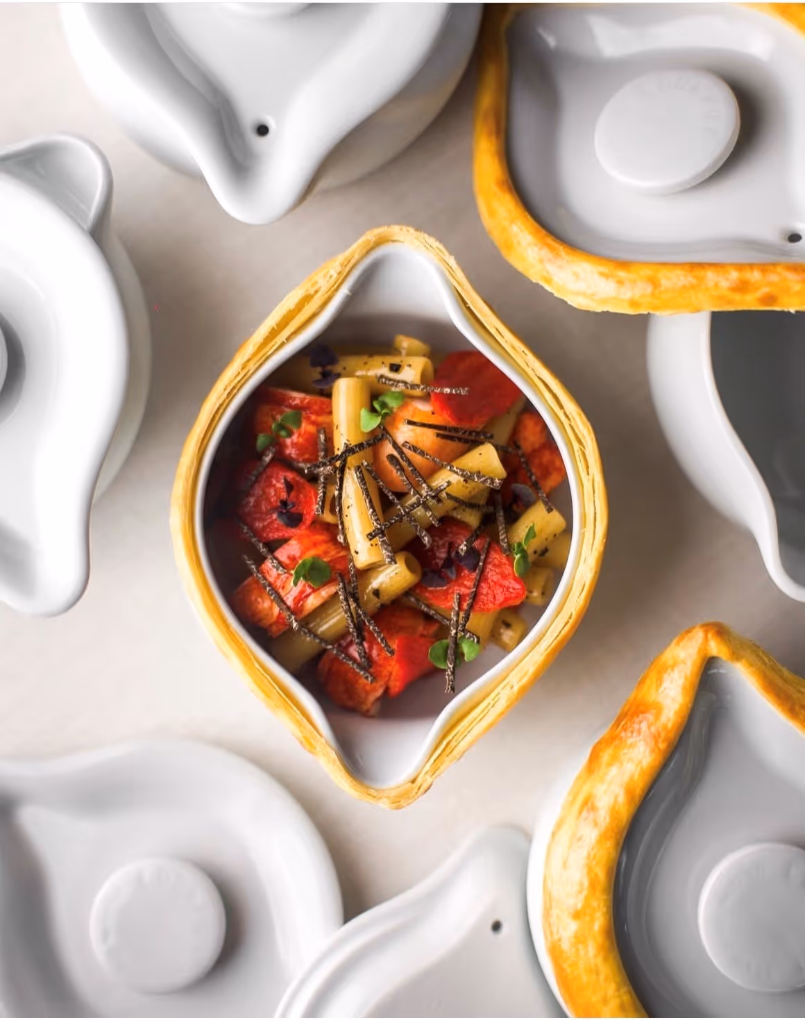 Plat gastronomique avec morceaux de légumes et herbes dans une porcelaine blanche Pillivuyt en forme de goutte avec une bordure dorée, entouré de plusieurs couvercles blancs.