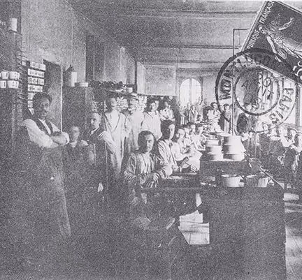 Photo historique de la manufacture de porcelaine Pillivuyt. Groupe d'ouvriers Pillivuyt posant dans une grande pièce d'usine avec des plateaux empilés sur une table.