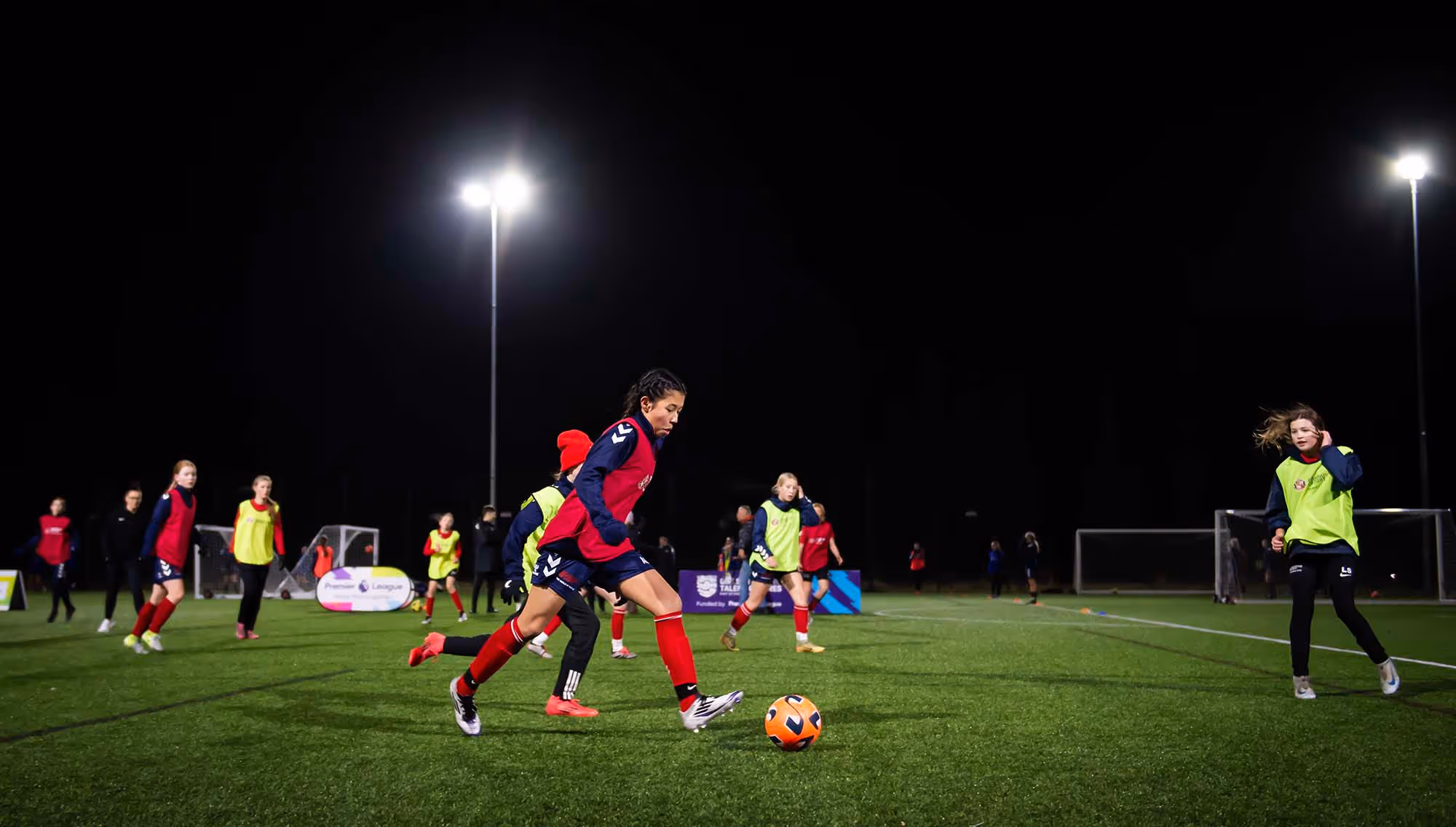 Players at an FA Emerging Talent Centre compete for the ball at night.