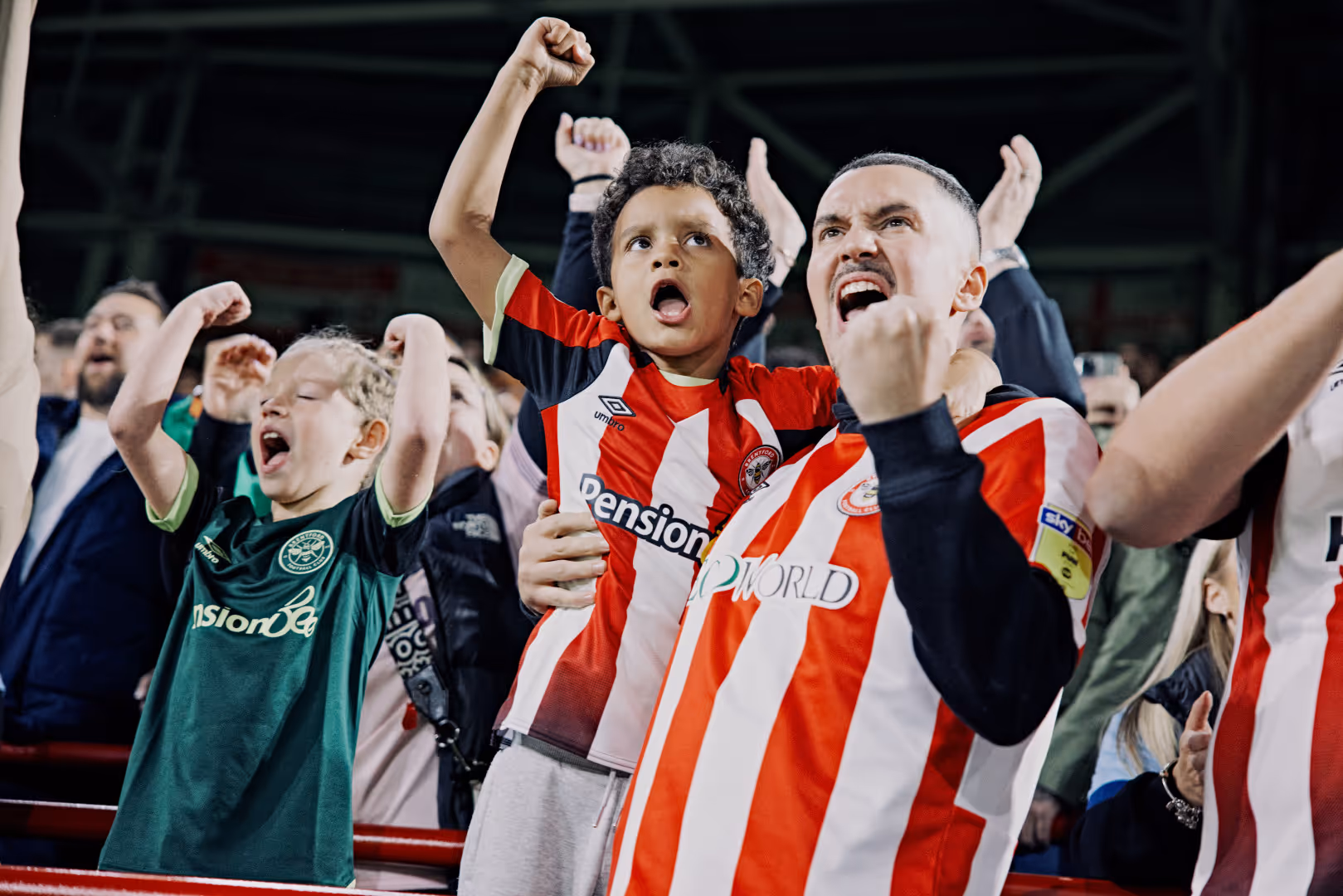 An adult and a child Brentford fan celebrate together, raising their fists. The adult holds the child in his arm.