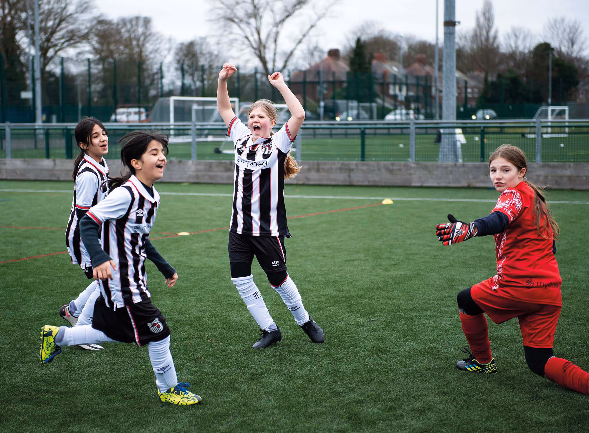 A group of girls celebrate on a football pitch while wearing black and white kits. A goalkeeper in red looks at the camera.