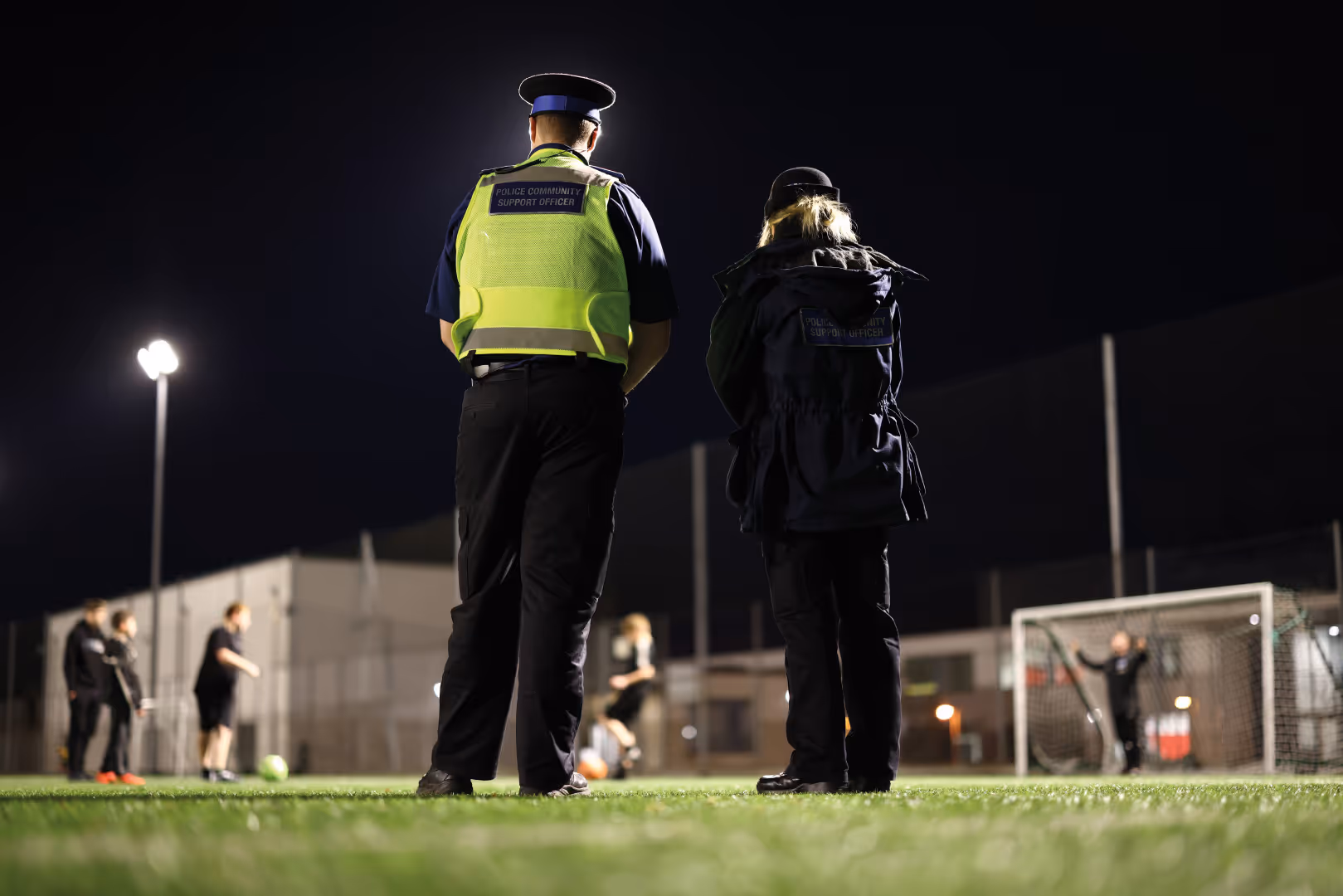 Two police officers stand with their backs to camera while watching community football.