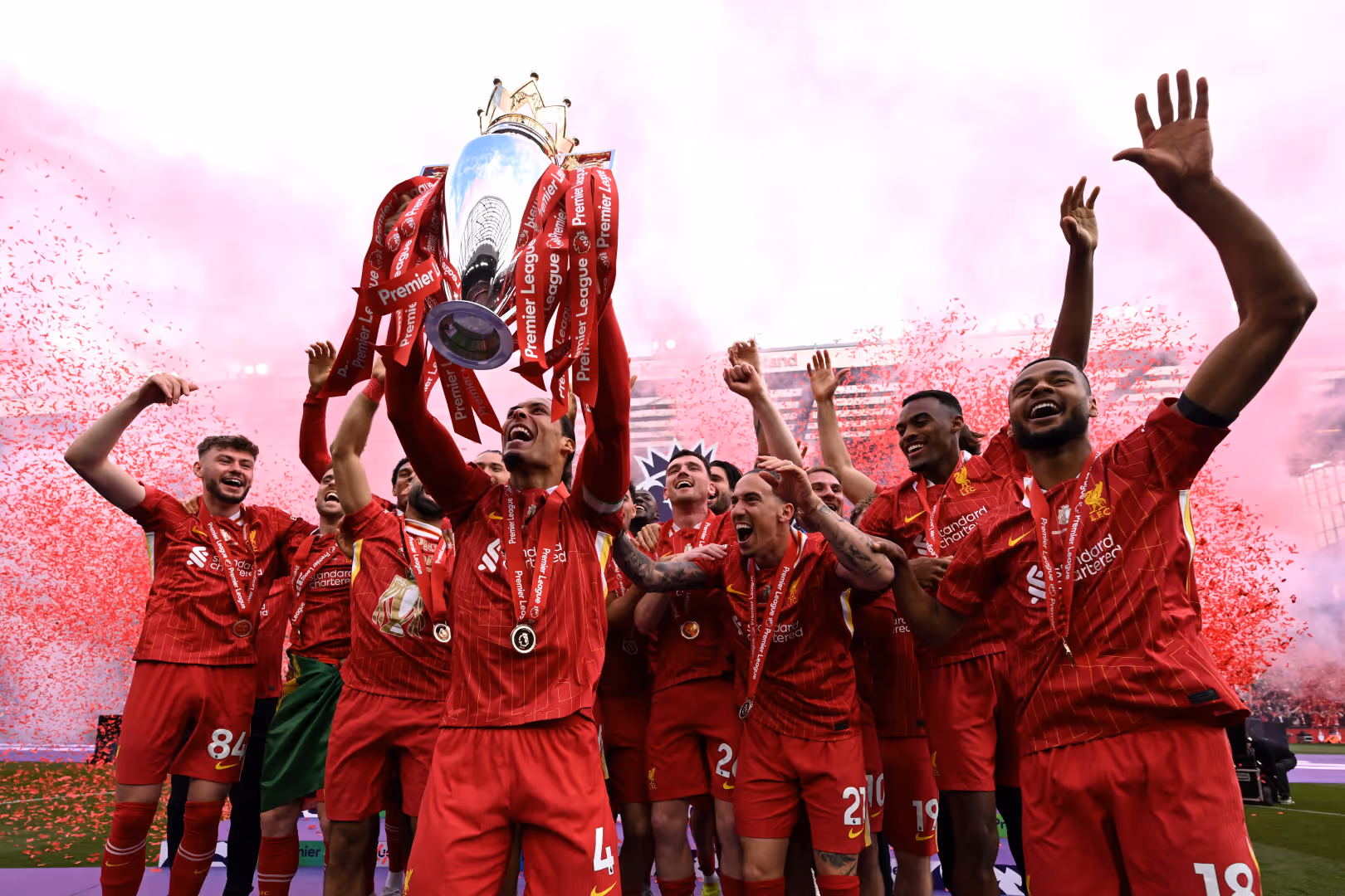 Liverpool players cheer and lift the Premier League Trophy as Champions. Red streamers are visible in the background.