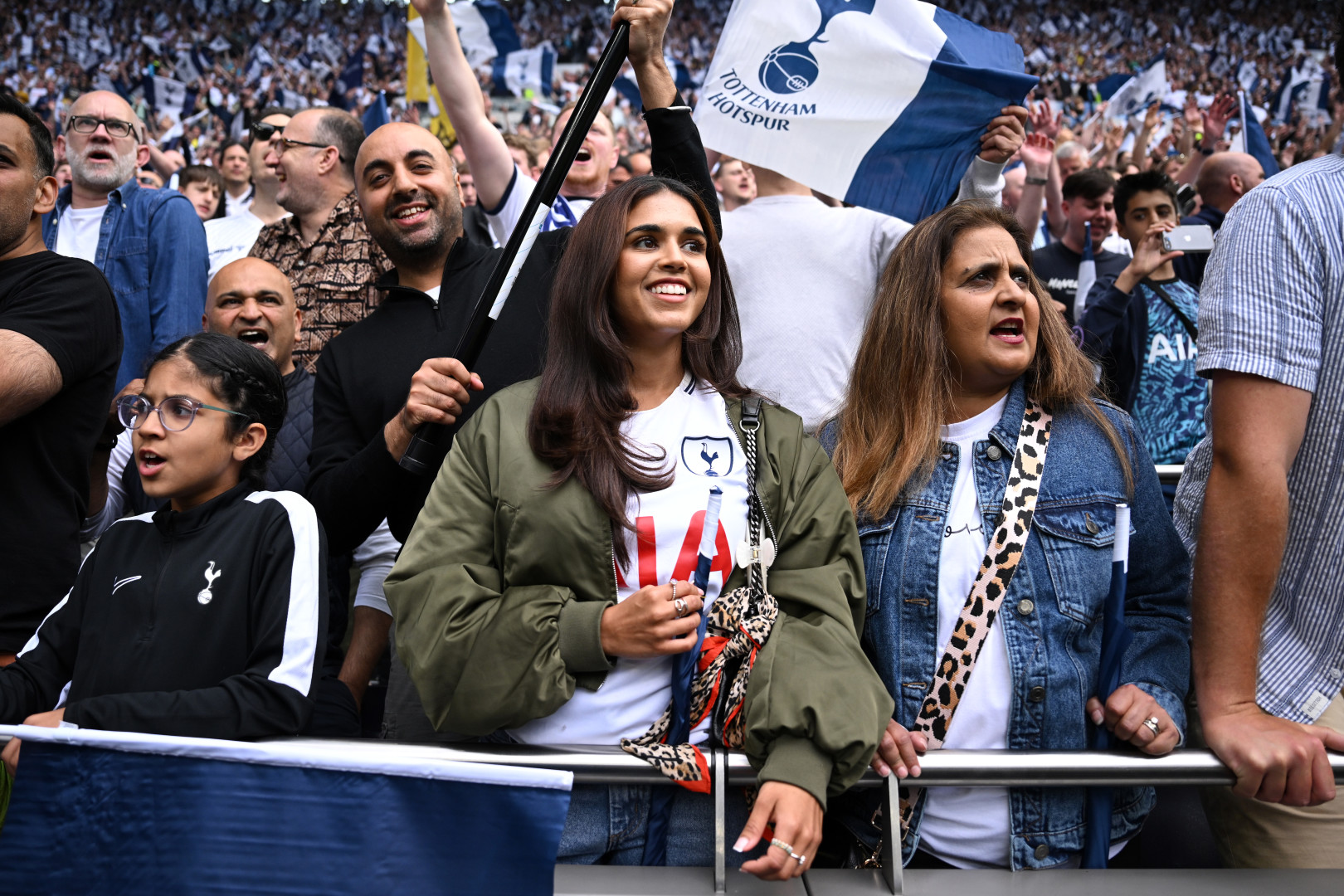 Tottenham Hotspur fans smile while looking out at the pitch and wearing Spurs shirts.