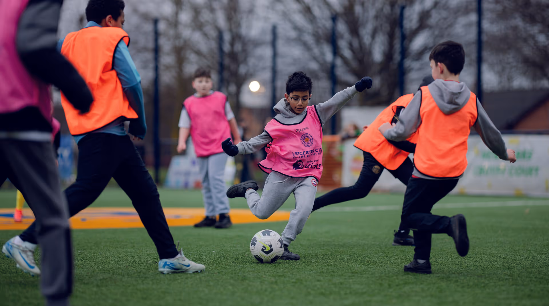 A Premier League Kicks participant strikes the ball while being challenged by another child. Both wear football bibs.