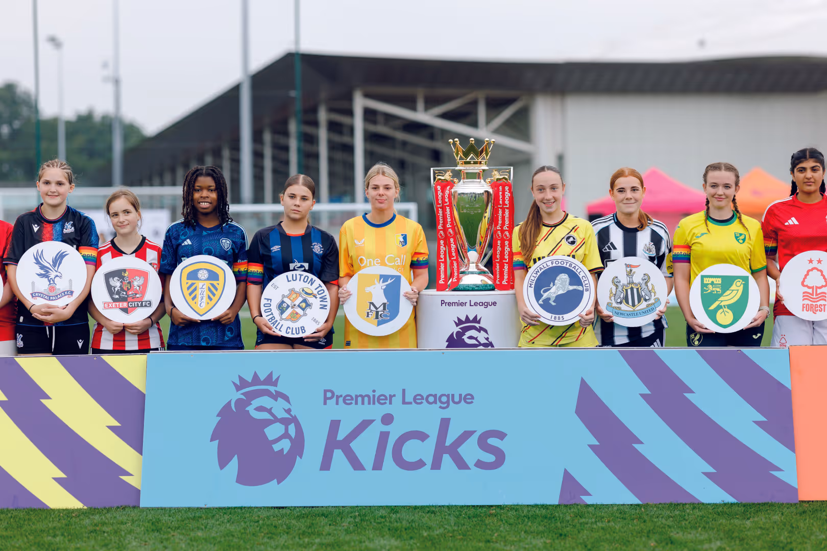 Female participants in the Premier League Kicks Cup hold their club crest and stand either side of the Premier League Trophy behind a blue board which has the Premier League Kicks logo on.