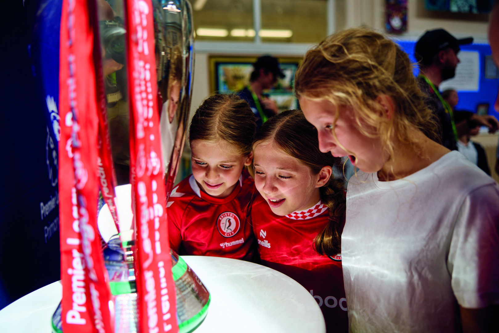 Young girls wearing Bristol City shirts smile while looking at the Premier League Trophy.