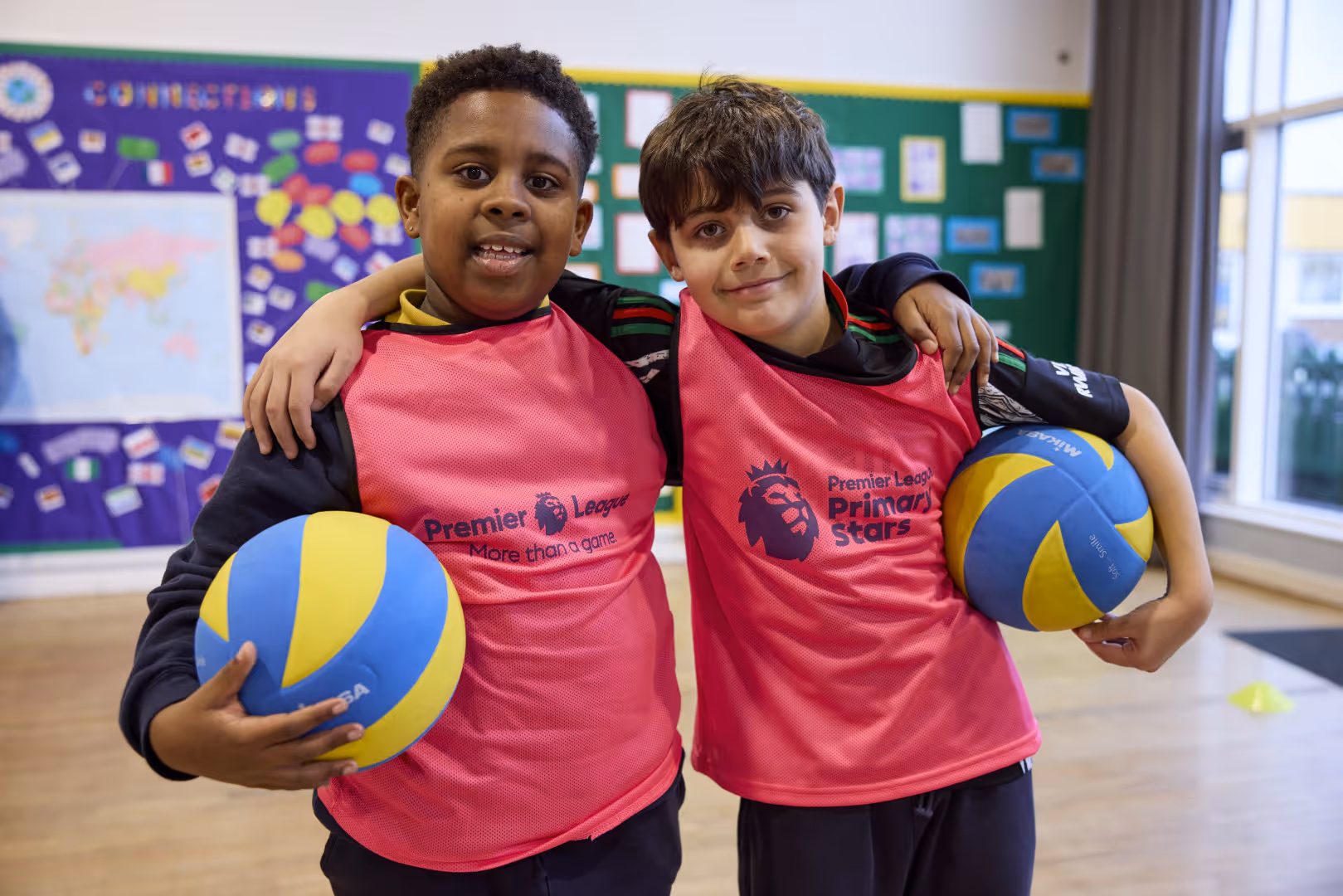 Two young boys smile with their arms around each other wearing pink Premier League bibs and holding balls.