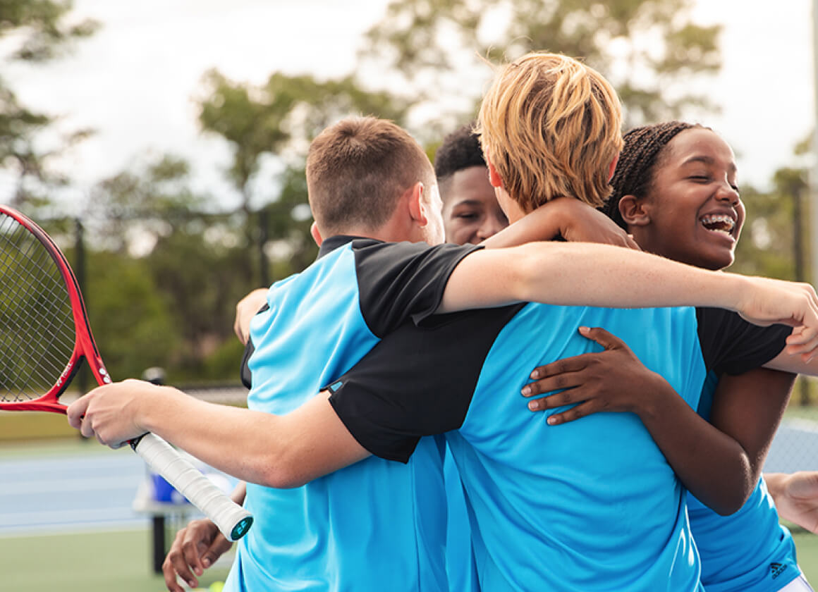Group of four young tennis players in blue uniforms joyfully hugging on a tennis court.
