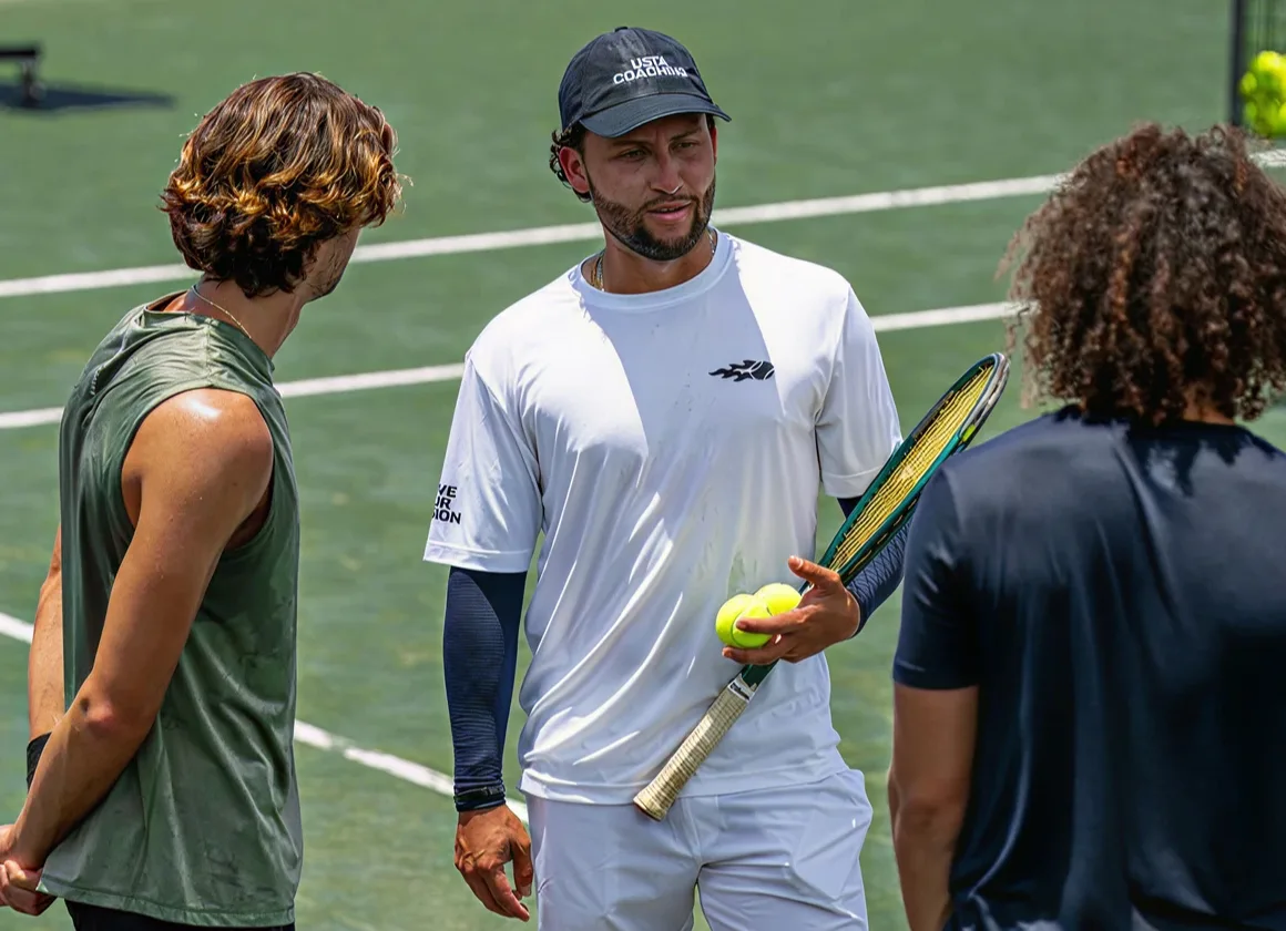 Tennis coach in white holding tennis balls and racket talking to two players on a court.