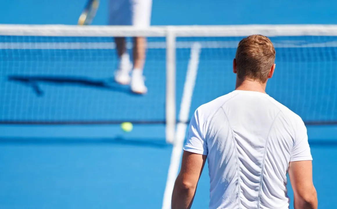 Man in white shirt standing on blue tennis court facing the net and another player on the opposite side.