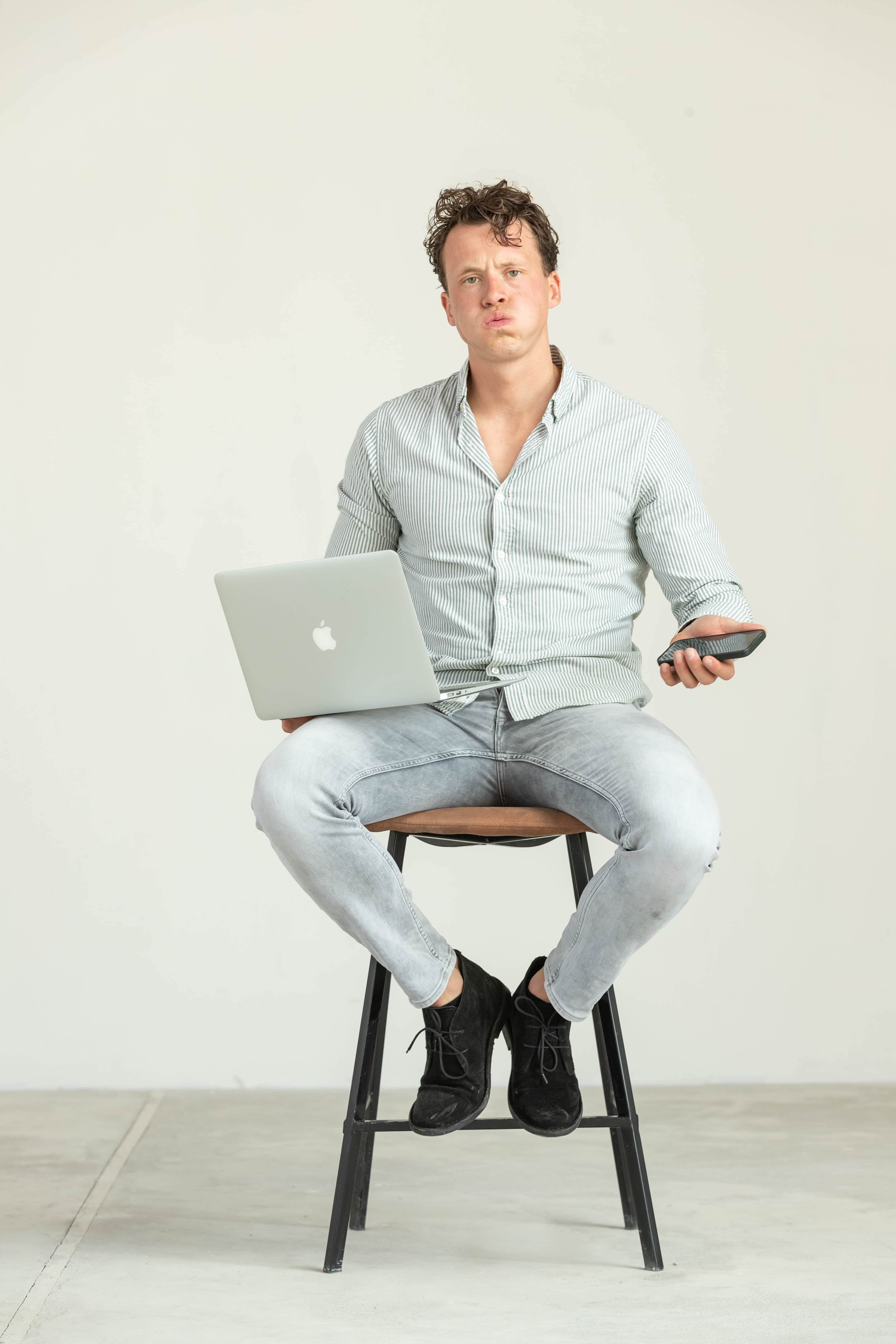 Man die met een laptop op schoot en een telefoon in zijn hand op een kruk zit in een lichte studio terwijl hij nadenkend voor zich uitkijkt