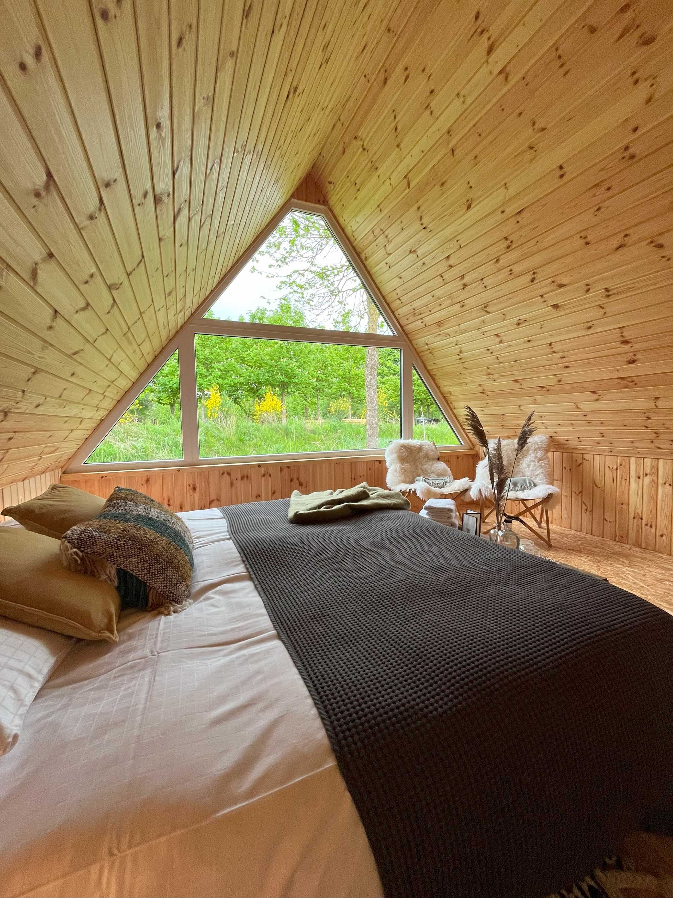 Soverom i en hytte med trekantet vindu som viser grønt landskap utendørs, en seng med brunt pledd og komfortable stoler med pelsdekor.