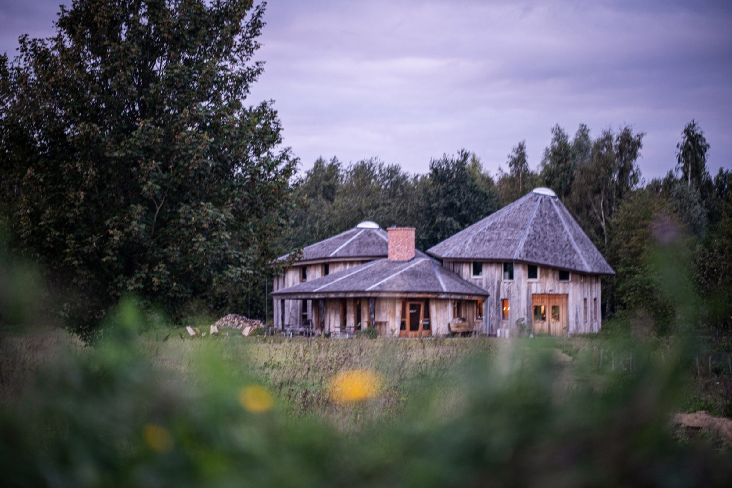 Tre rundt trebuer med trekledning midt i en grønn eng omgitt av trær under en lilafarget himmel.