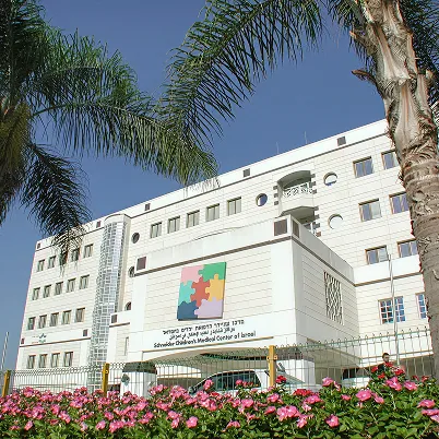 Schneider Children's Medical Center of Israel building with white exterior, colorful puzzle logo, palm trees, and pink flowers in front under clear blue sky.