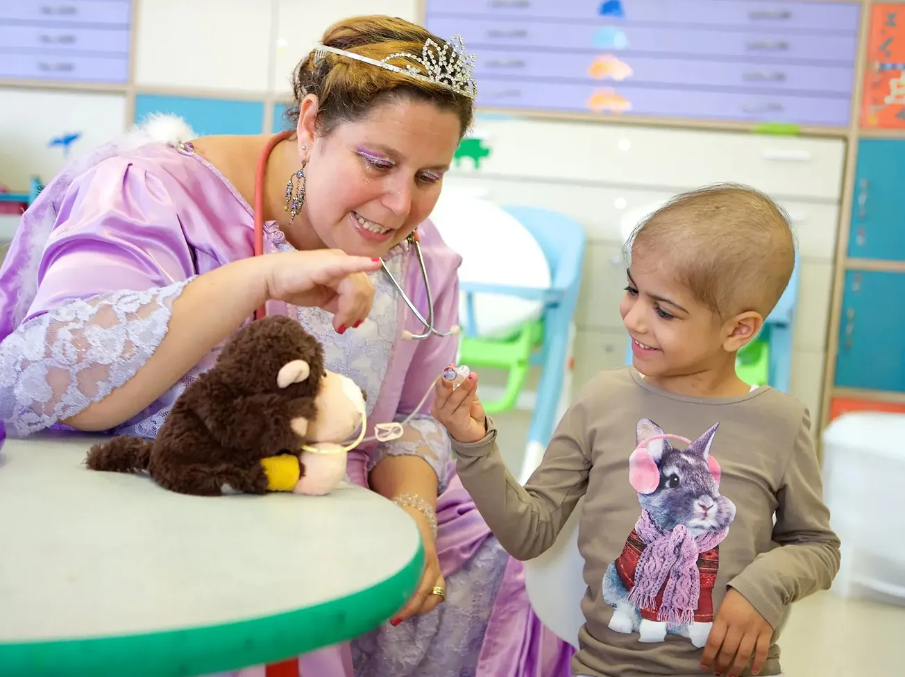 Smiling child with no hair holds a small medical toy while a woman in a purple dress and tiara engages with a stuffed monkey toy on a table.
