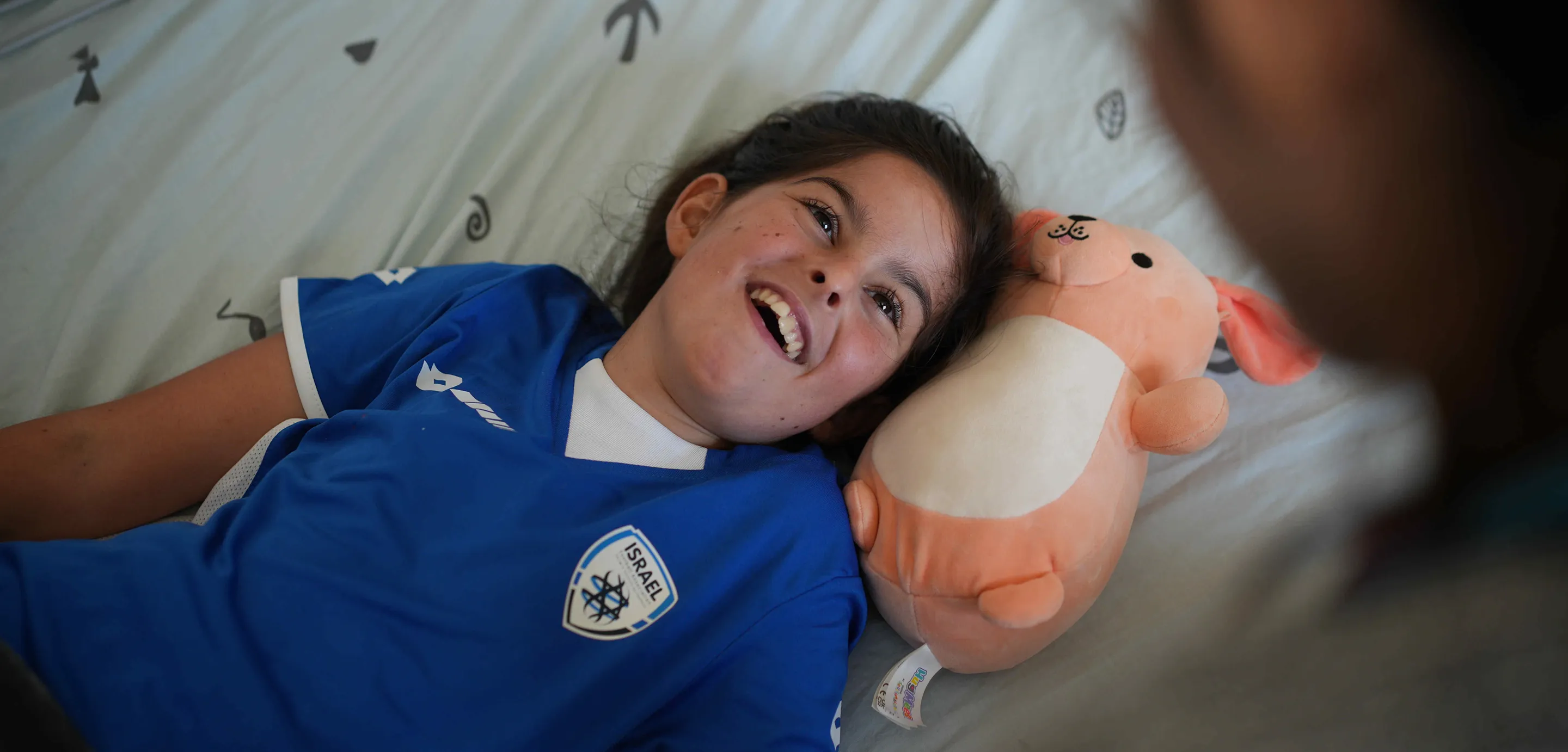 Smiling girl lying on a bed with her head resting on a pink stuffed animal resembling a bear or dog.