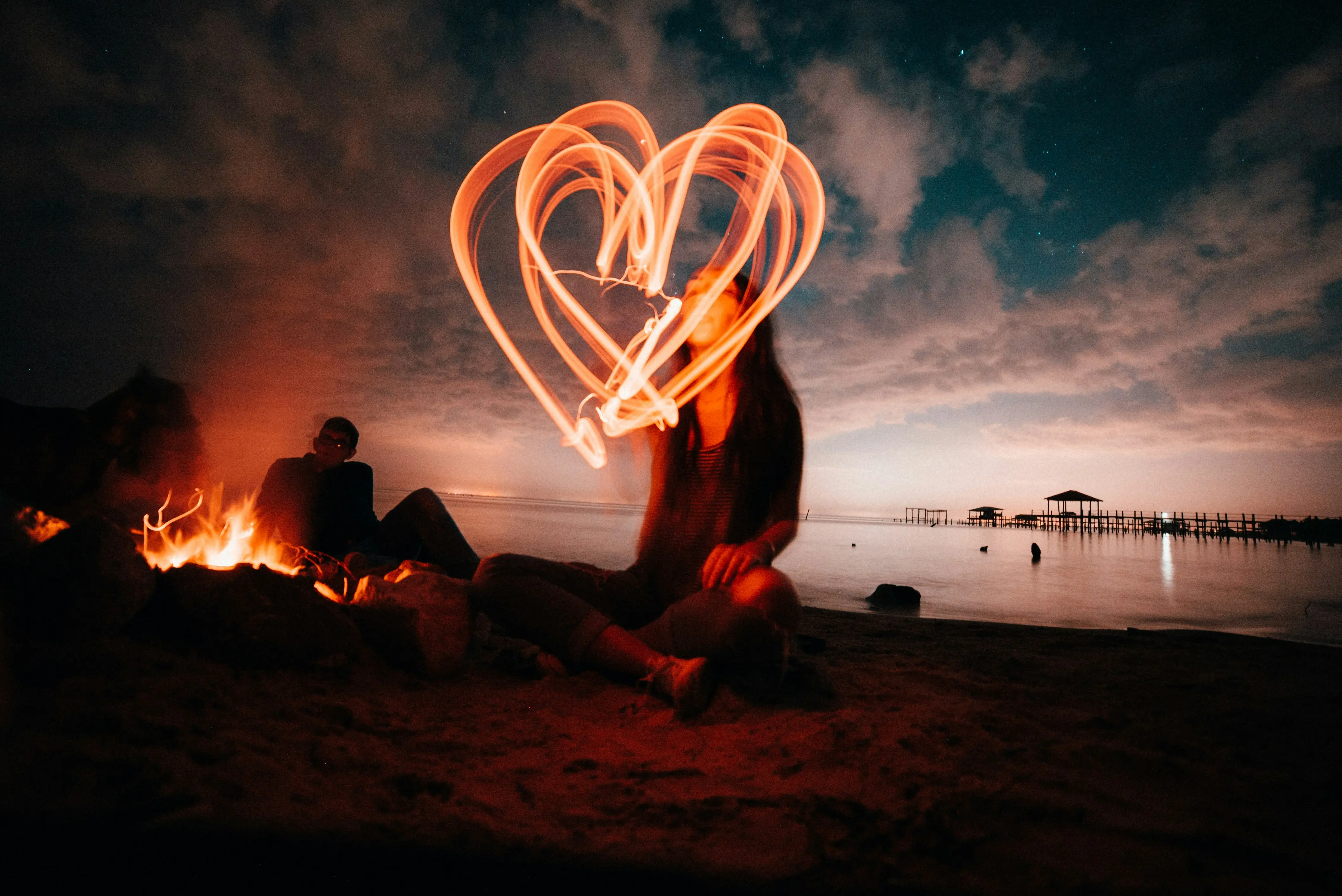 Person assise sur une plage la nuit dessinant un grand cœur lumineux avec une source de lumière. Un feu de camp éclaire deux autres personnes, un ponton est visible sur l'eau calme.