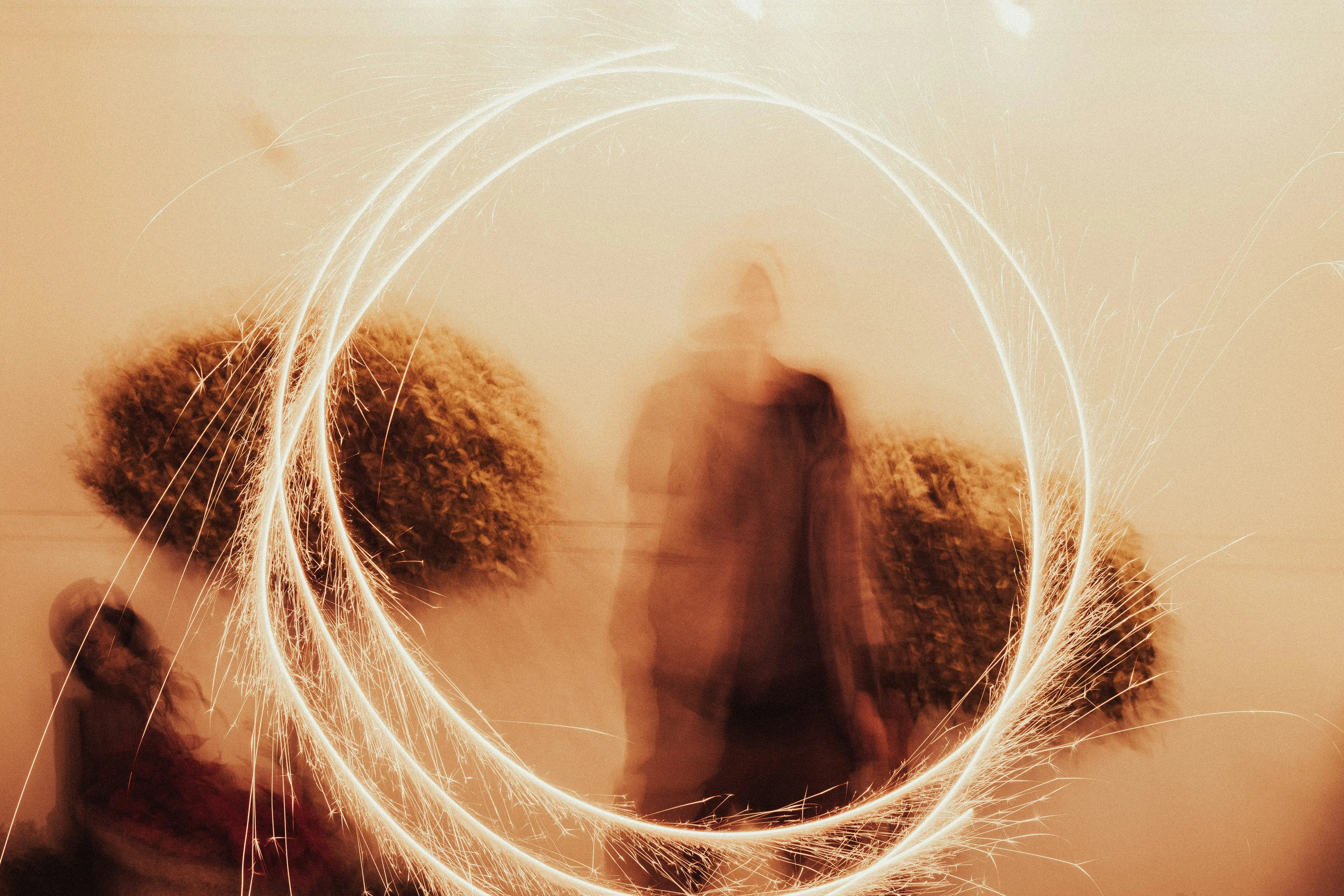 Une silhouette floue entourée d'anneaux lumineux scintillants créés par des étincelles en mouvement dans un décor extérieur avec des buissons.