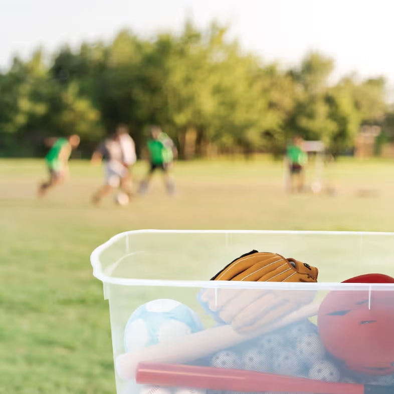 Plastic bin filled with sports equipment including a baseball glove, baseball bat, ball, and bowling ball with players in green jerseys blurred in the background on a grassy field.