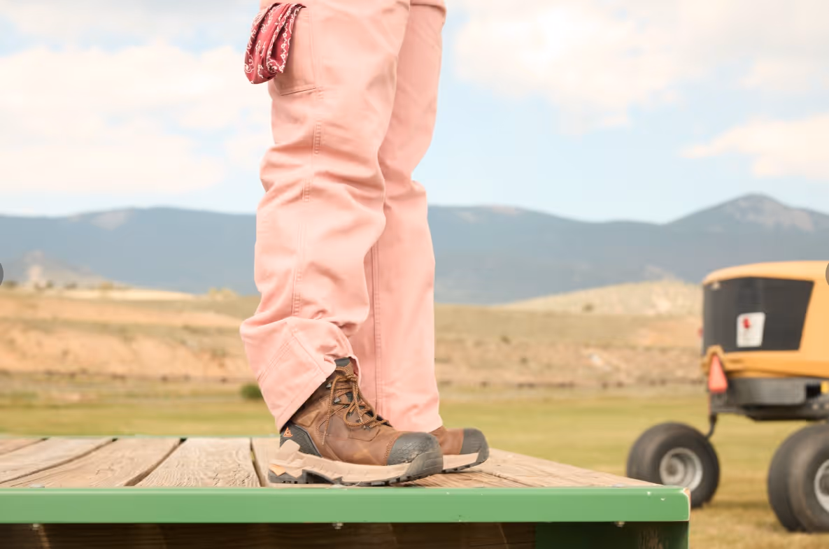 Person wearing brown work boots and pink pants standing on a wooden platform outdoors with mountains in the background.