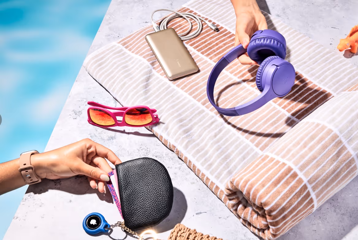 Two hands near poolside holding a black leather pouch and purple wireless headphones on a striped towel beside pink sunglasses and a gold power bank.
