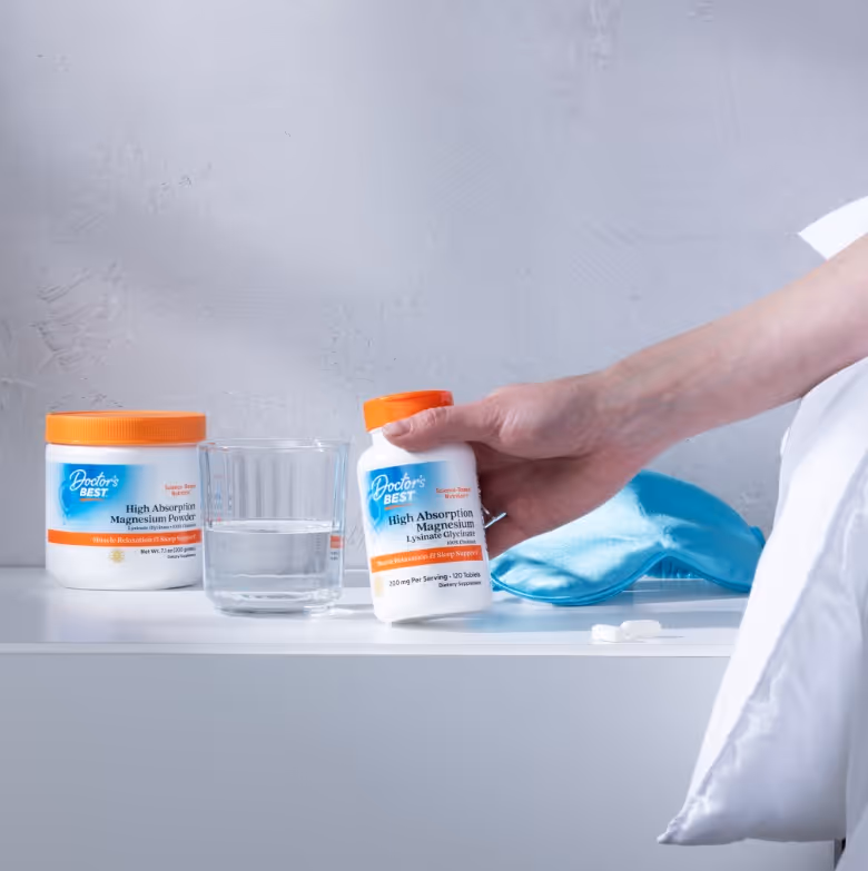 Hand holding a bottle of Doctor's Best High Absorption Magnesium supplements next to a glass of water, magnesium powder container, and a blue sleep mask on a white surface.