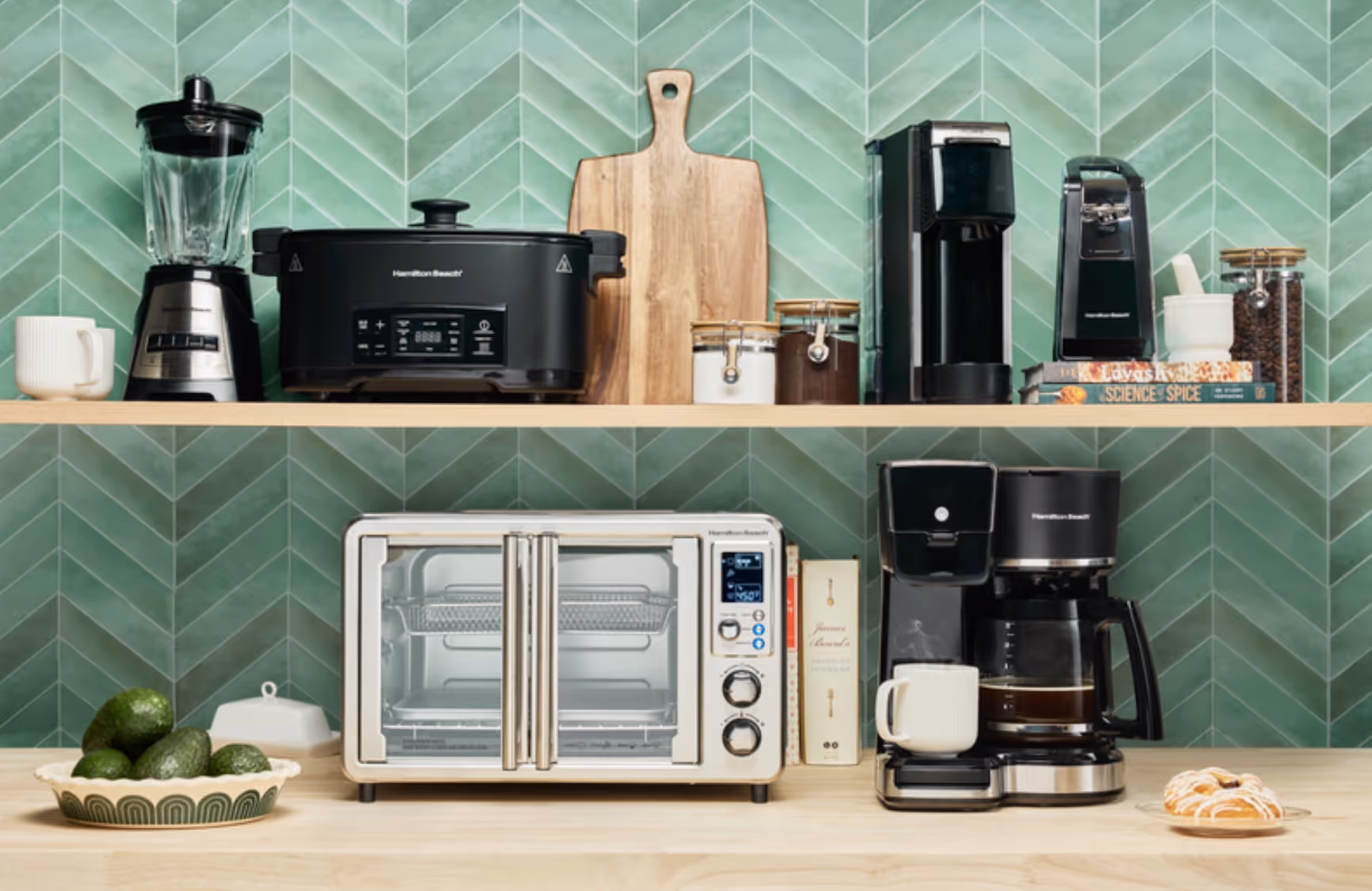 Kitchen countertop and shelf displaying a blender, slow cooker, cutting board, jars, coffee maker, toaster oven, avocados in a bowl, and a glazed pastry on a plate.