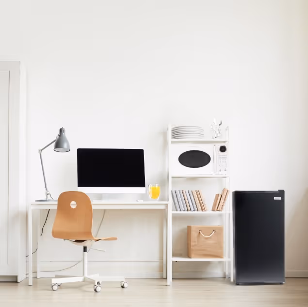Minimalist home office setup with a wooden chair, white desk holding a computer and lamp, adjacent white shelving with microwave, books, plates, and a small black refrigerator.