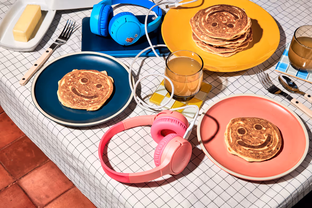 Table with smiling face pancakes on colorful plates, glasses of juice, butter dish, and two sets of headphones connected by a white cable.