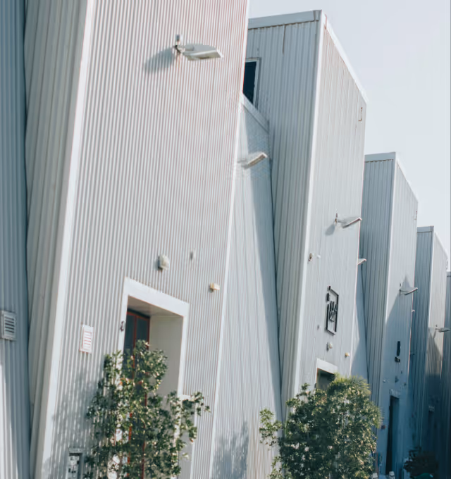 Row of modern industrial buildings with tall, slanted white corrugated metal walls and small trees in front.