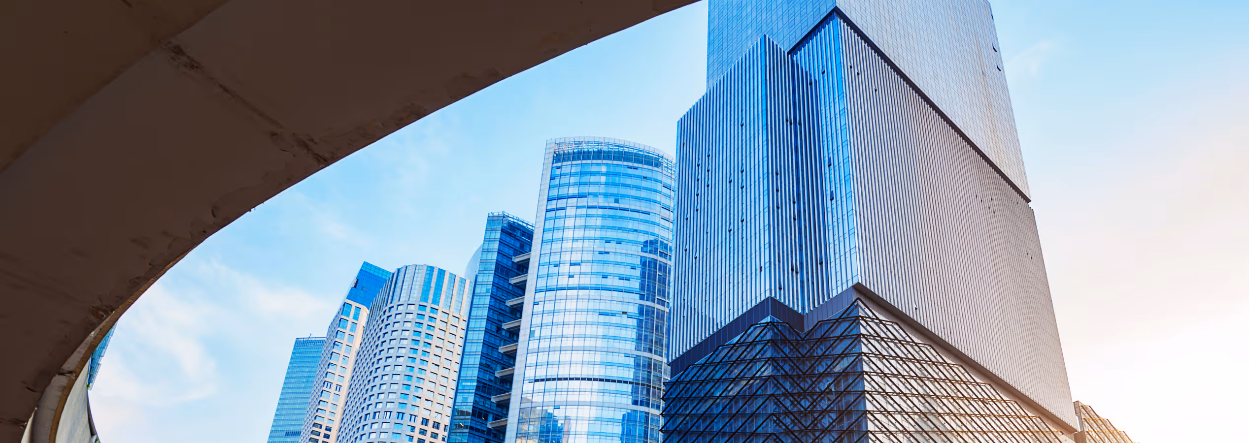 Modern city skyline viewed through a large curved architectural frame with greenery and tiled plaza in the foreground.