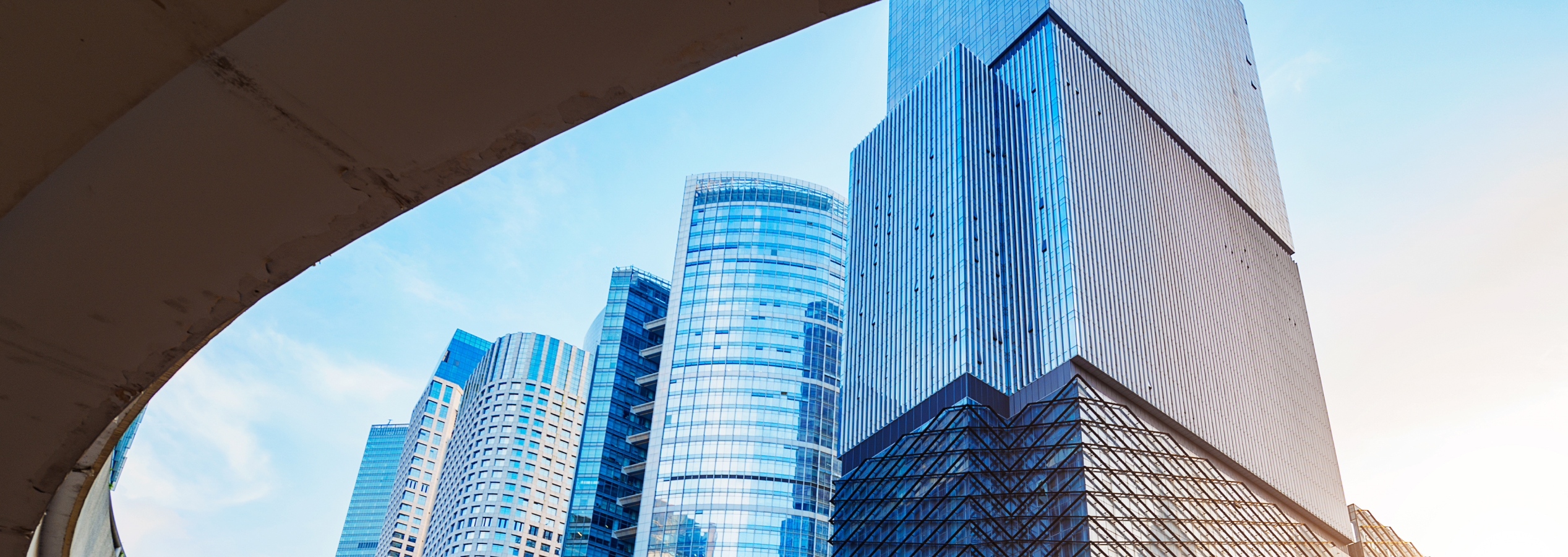 Modern city skyline viewed through a large curved architectural frame with greenery and tiled plaza in the foreground.