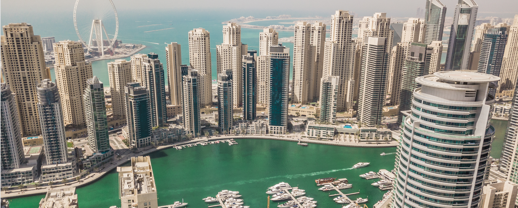 Aerial view of a marina with numerous yachts docked, surrounded by tall modern buildings and a large ferris wheel near the coastline.