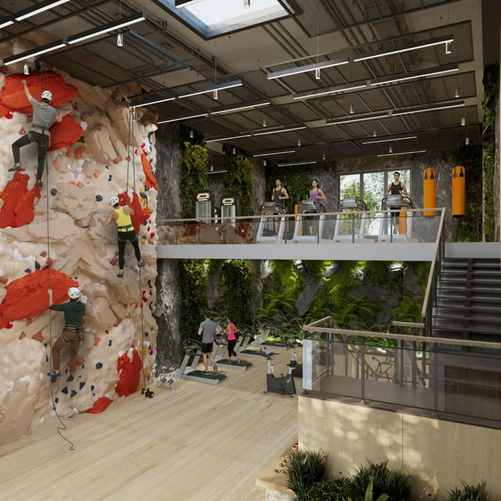 Climbers on an indoor climbing wall beside a gym with treadmills on a planted mezzanine.