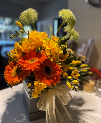 Bright orange and yellow flowers arranged in a gray gift box with a white ribbon bow.