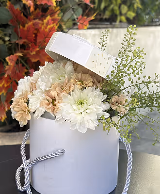 White round gift box with a lid leaning open, containing white and light peach flowers with green foliage.