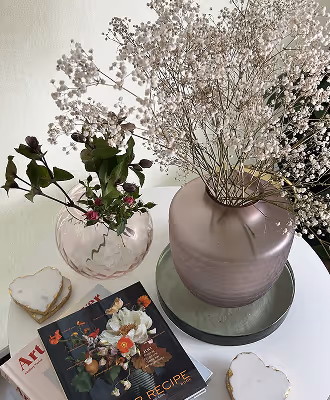 Two vases on a white table, one with delicate baby's breath flowers and the other with green leaves and small red flowers, alongside magazines and heart-shaped coasters.