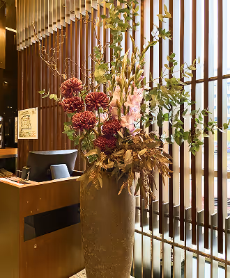 Tall floral arrangement with red and pink flowers and green foliage in a large textured vase near wooden slatted window panels.