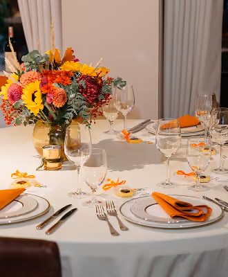Round table with floral centerpiece, orange napkins, and multiple wine glasses set for a formal event.