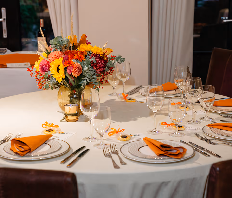 Round table set with white cloth, orange napkins, glassware, and a colorful flower centerpiece.