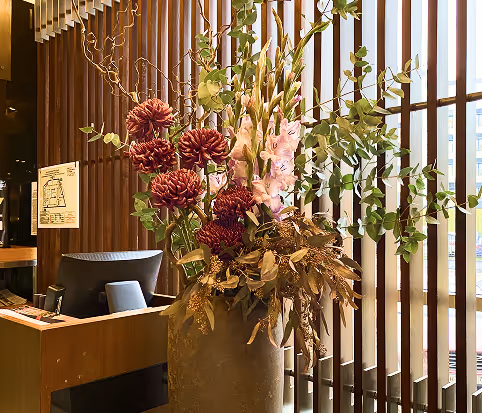 Large floral arrangement with burgundy, pink flowers, and green foliage in a tall vase by a wooden slatted window near a desk.