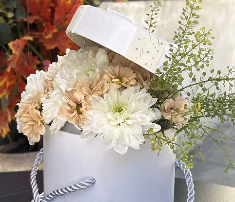 White and peach flowers arranged with greenery in an open white gift box with a rope handle.