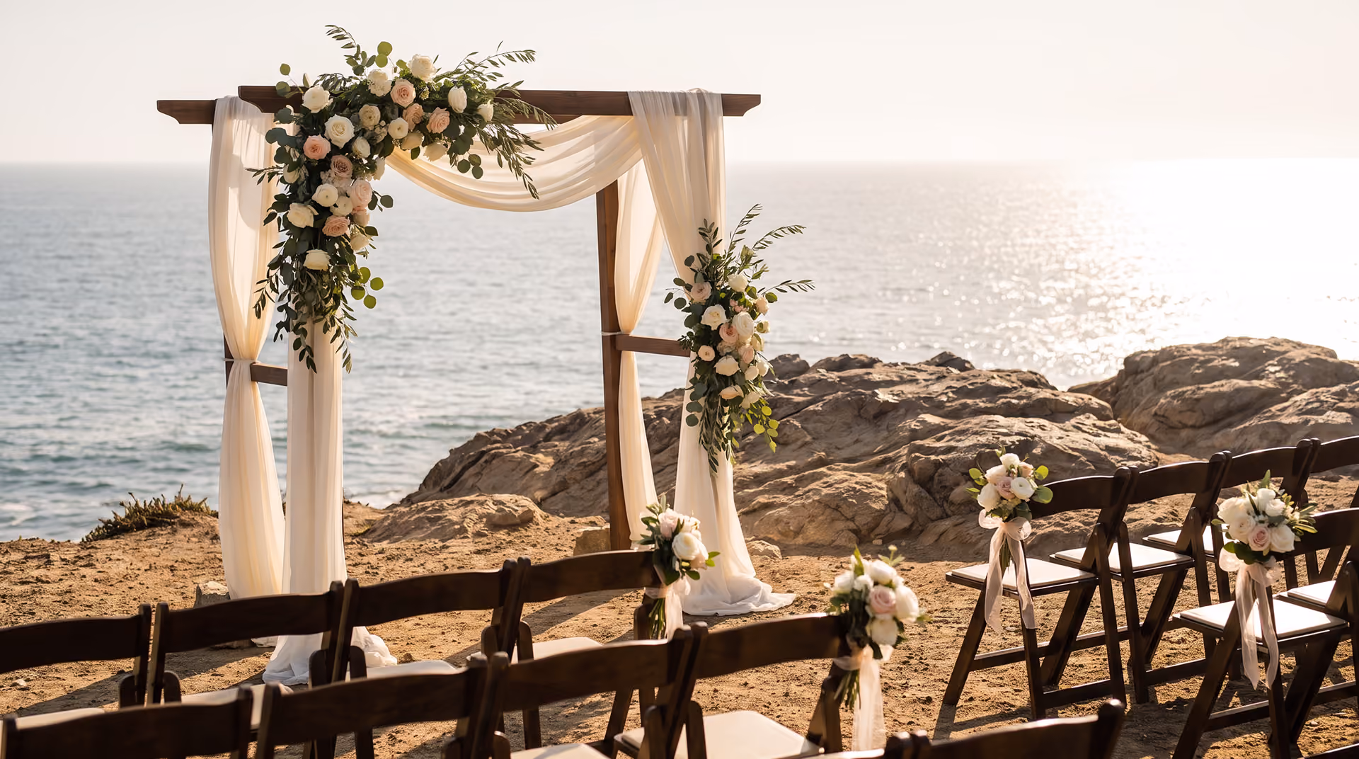 Wooden wedding arch with white drapes and flowers on a rocky beach overlooking the ocean at sunset.