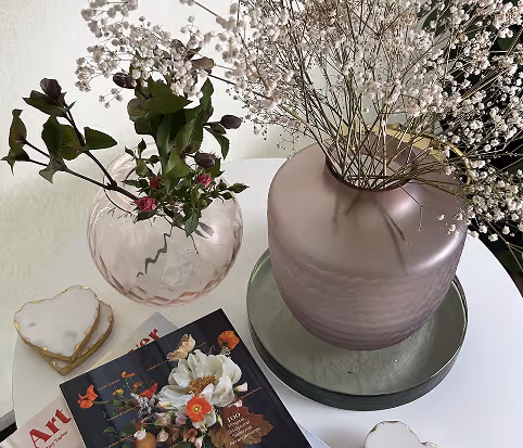 Two vases on a white table, one pink frosted with baby's breath flowers and one clear pink with green leaves and small red buds, beside an art magazine and heart-shaped items.
