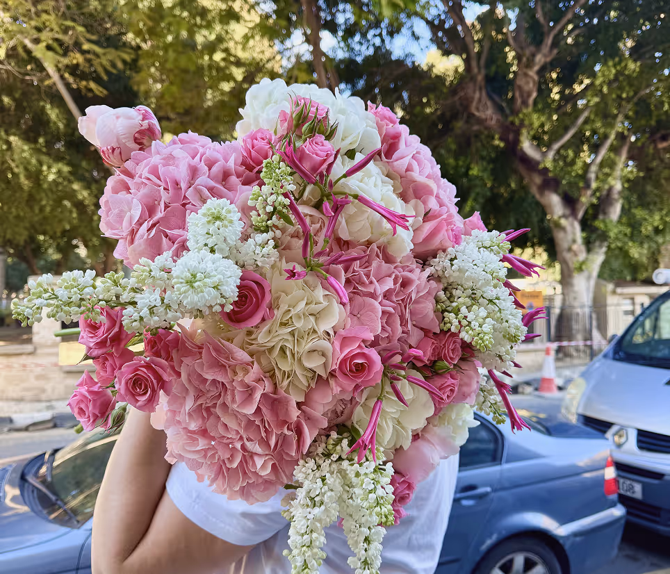 Person holding a large bouquet of pink and white flowers outdoors with cars and trees in background.