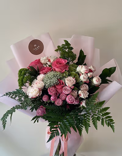 Bouquet of pink and white roses with green foliage wrapped in light pink paper and tied with a pink ribbon.