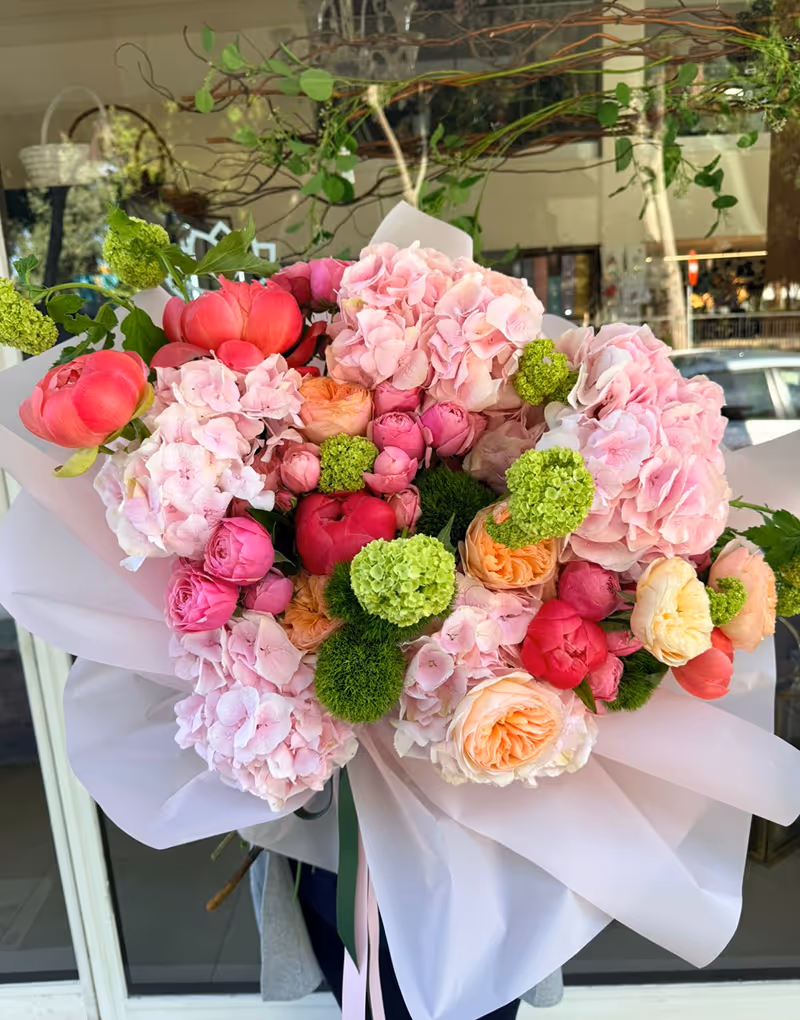 Bouquet of pink hydrangeas, green flowers, and assorted orange and red blooms wrapped in white paper.