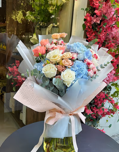 Bouquet of mixed flowers including white roses, pink and peach roses, and blue hydrangeas wrapped in light pink and white paper with a ribbon on a black table.