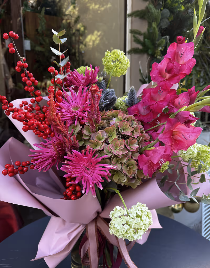 Bouquet with bright pink flowers, green hydrangeas, red berries, and foliage wrapped in pink paper.